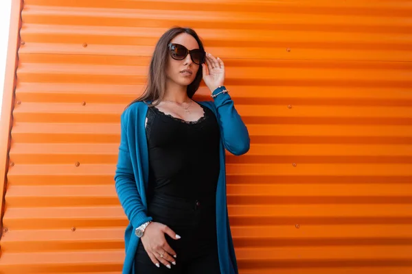 Pretty young urban hipster woman in a knitted blue cape in stylish sunglasses in a fashionable black t-shirt is standing near the metallic orange wall outdoors. Beautiful girl model in the city. Style