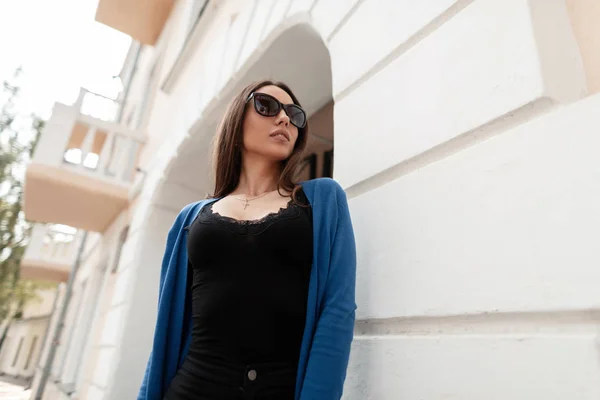 Roztomilá mladá žena s hipíky v tmavých slunečních brýlích ve stylově modrém pleteném plášti v džínách, které stojí nedaleko budovy v letním dnu. Krásná dívka si užívá víkend. — Stock fotografie