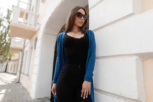 Bella giovane hipster donna in elegante top nero in un mantello a maglia in jeans alla moda in occhiali da sole scuri sta posando vicino a un edificio moderno della città. Attraente ragazza europea all'aperto. Stile estivo . — Foto Stock