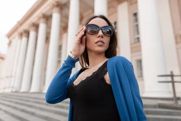 Αρκετά ελκυστική νεαρή γυναίκα χίππις με σέξι χείλη σε κομψά γυαλιά ηλίου σε ένα μοντέρνο μπλε πλεκτό Ακρωτήριο σε μια μοντέρνα μαύρη δαντέλα κορυφή βόλτες κοντά στο κτίριο με κολώνες. Όμορφο μοντέλο κορίτσι. — Φωτογραφία Αρχείου