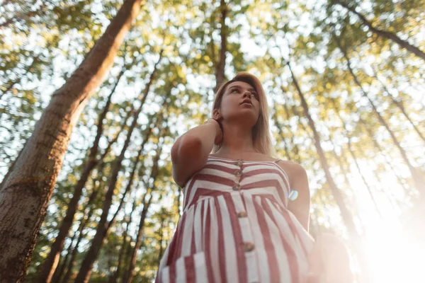 Pohled na krásnou mladou blonďatou ženu ve stylově růžově pruhované sluneční košili v lese mezi zelenými stromy za jasného slunného letního dne. Atraktivní dívka relaxuje v přírodě a požívá slunce. — Stock fotografie