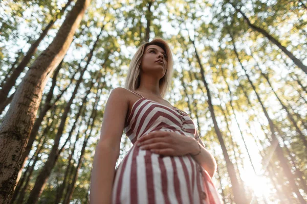 Attraktiv ung kvinna blondin i en sommar snygg randig sundress vilar i skogen på bakgrunden av starkt solljus. Vacker flicka njuter av solen utomhus. Mode kvinnor klä. Nedersta vyn. — Stockfoto