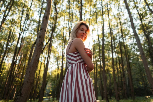 Attraktive glückliche junge Frau in einem stilvollen rosa gestreiften Kleid mit einem süßen Lächeln entspannt im Freien. fröhliche Mädchen ruhen sich zwischen den Bäumen im Wald aus und genießen die strahlende Sommersonne. — Stockfoto