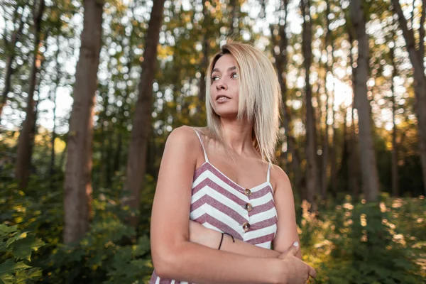 Całkiem atrakcyjna młoda kobieta z blond włosami z niebieskimi pięknymi oczami ze słodkim uśmiechem w modnej letniej sukni w paski sundress w lesie w słoneczny dzień. Piękna dziewczyna model relaksuje na zewnątrz. — Zdjęcie stockowe