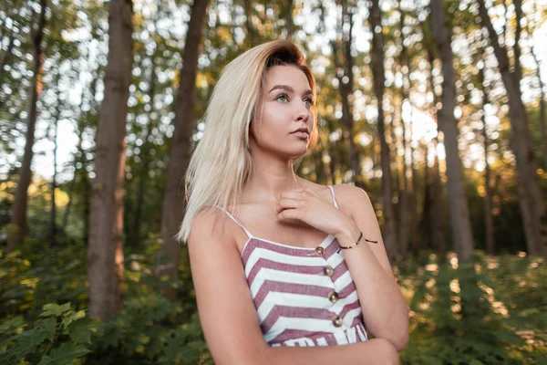 Całkiem seksowna młoda blond kobieta w stylowej różowy pasiasty sundress stwarzające w słoneczny dzień wśród drzew w lesie. Modny model zmysłowej dziewczyny odpoczywa w przyrodzie. Słoneczny letni dzień. — Zdjęcie stockowe