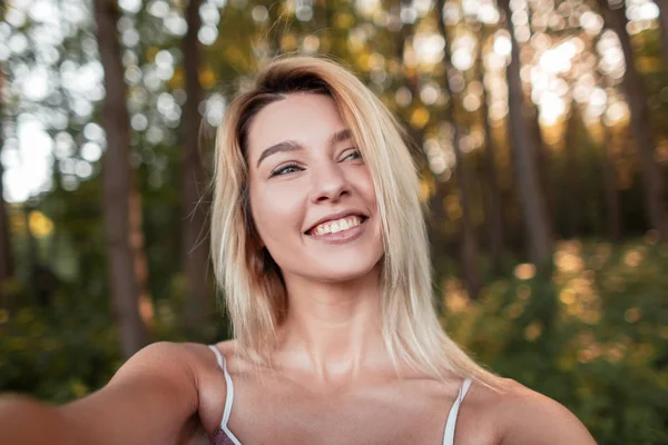 Ziemlich positive junge blonde Frau mit einem schönen Lächeln mit blauen Augen macht ein Selfie im Wald an einem sonnigen Sommertag. fröhliche Mädchen entspannen sich gerne in der Natur. — Stockfoto