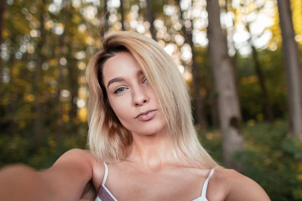 Jonge stijlvolle blonde vrouw in een gestreepte roze modieuze sundress maakt selfie staande in het bos op een heldere zonnige zomerdag. Schattig meisje met blauwe ogen is het nemen van een foto van zichzelf. — Stockfoto