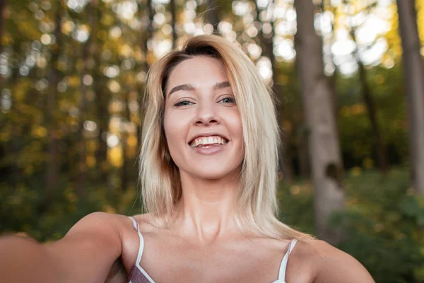 Glückliche junge blonde Frau mit positivem Lächeln in einem trendigen rosa Kleid macht ein Selfie auf dem Hintergrund eines Baumes und eines Sonnenuntergangs im Wald. hübsches fröhliches Mädchen, das an einem Sommertag in der Natur ruht. — Stockfoto