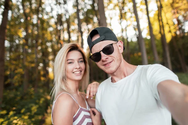 Stijlvolle knappe jonge man met een gelukkige vrouw in modieuze zomer kleren met glazen in een GLB wandelingen in het bos en het nemen van selfies. Gelukkige vrouw met een mooie man die foto's van zichzelf neemt — Stockfoto