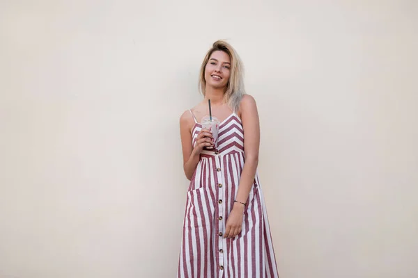 A jovem mulher positiva moderna em um vestido listrado rosa longo elegante fica perto de uma parede vintage com um saboroso milkshake nas mãos. Menina feliz loira com um sorriso bonito ao ar livre. Estilo de verão . — Fotografia de Stock