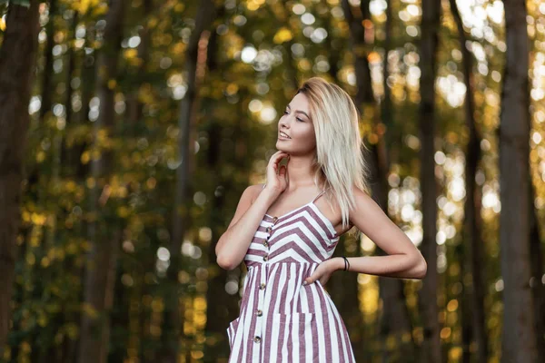 Efektowna Ładna natura młoda kobieta ze słodkim uśmiechem w modnym różowym stroju w paski relaksuje stojąc w lesie w słoneczny letni dzień. Atrakcyjna dziewczyna cieszy się ciepłym słońcem w pobliżu drzew — Zdjęcie stockowe