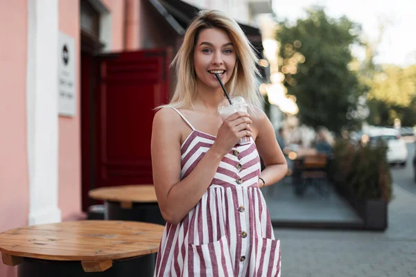 Забавная веселая молодая блондинка в модном розовом полосатом платье стоит в городе и пьет молочный сладкий коктейль. Симпатичная счастливая девушка наслаждается прогулкой в солнечный летний день . — стоковое фото