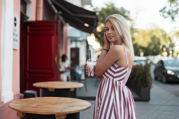 Urban giovane donna carina in un elegante lungo rosa a righe prendisole stand con un gustoso milkshake in mano. Bella ragazza bionda con un dolce sorriso in città all'aperto. Moda estate . — Foto Stock