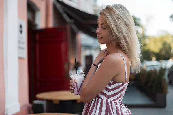 Mujer rubia joven hermosa bastante moderna en un elegante vestido de rayas rosadas con un paseo de bebida de leche fría dulce en la calle cerca de un café abierto. Linda chica modelo disfruta al aire libre a pie. Buen día de verano . —  Fotos de Stock