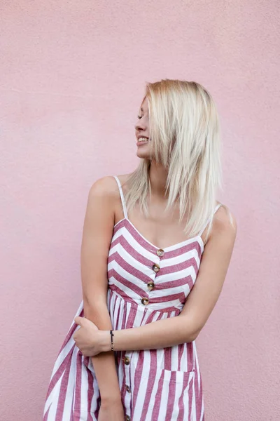 Linda mujer joven y elegante rubia en vestido a rayas de moda posando cerca de una pared vintage rosa en la calle en la ciudad. Hermosa chica glamorosa alegre se relaja al aire libre . —  Fotos de Stock