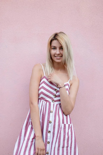 Mujer rubia bonita y divertida en un vestido a rayas rosa de moda con una sonrisa positiva posa cerca de la pared rosa vintage al aire libre en la ciudad en un día de verano. Linda chica alegre disfruta de un paseo . —  Fotos de Stock