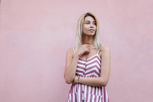 Atractiva joven rubia en un verano elegante vestido de rayas descansa cerca de una pared vintage rosa en la ciudad. Hermosa chica linda modelo al aire libre. Moda ropa de mujer . — Foto de Stock