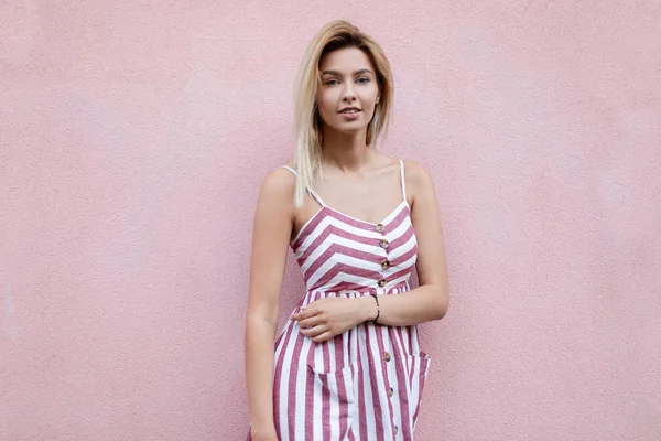 American glamorous young woman with blond hair in a stylish striped pink dress posing standing near a pink vintage building on a warm summer day outdoors. Fashionable attractive girl model.Youth style — Stock Photo, Image
