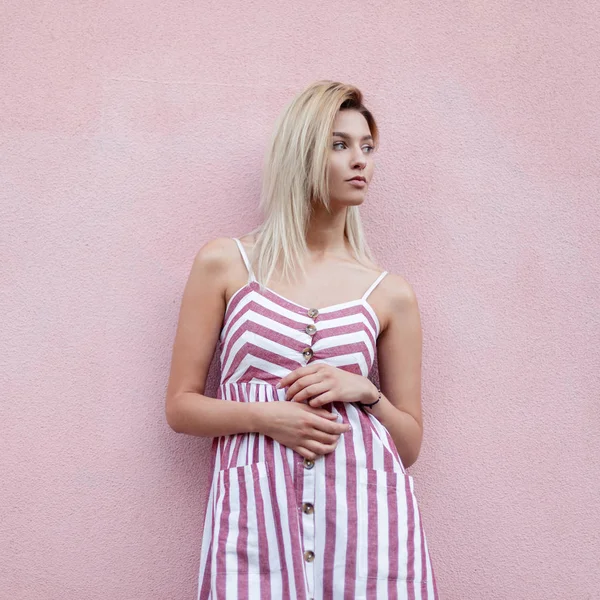 Modische hübsche junge Frau Blondine in trendigen rosa gestreiften Kleid posiert in der Nähe einer rosa Vintage-Wand auf der Straße in der Stadt. schöne glamouröse freudige Mädchen entspannt sich im Freien. Sommerkleider für Frauen. — Stockfoto