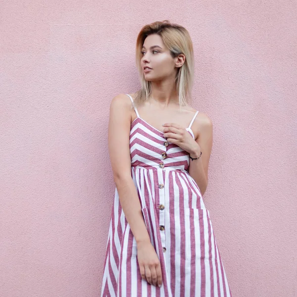 Mujer rubia bonita europea en un verano elegante vestido de rayas descansa cerca de una pared vintage rosa en la ciudad. Hermosa chica urbana modelo al aire libre. Ropa de mujer de moda. Estilo de verano . —  Fotos de Stock