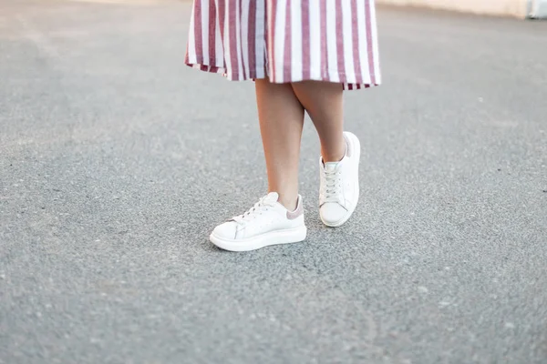 Жіночі ноги в стильній рожевій довгій сукні в модних білих шкіряних кросівок стоять на тротуарі. Молода жінка йде по вулиці в літній день. Нова колекція модного сучасного взуття . — стокове фото