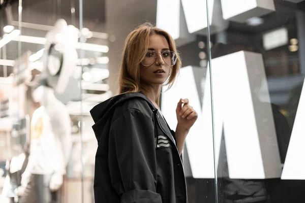 Hermosa joven hipster pelirroja en chaqueta con capucha con estilo en gafas de moda está de pie en una tienda cerca de la pared de vidrio brillante. Chica urbana moderna disfruta de un paseo por el centro comercial. Estilo . — Foto de Stock