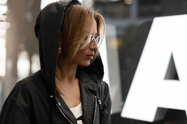 Sexy jeune femme aux cheveux roux hipster dans des lunettes élégantes dans une veste à capuche à la mode est debout dans un centre commercial près d'une vitrine en verre moderne lumineux. Belle fille en vacances. Style jeunesse . — Photo