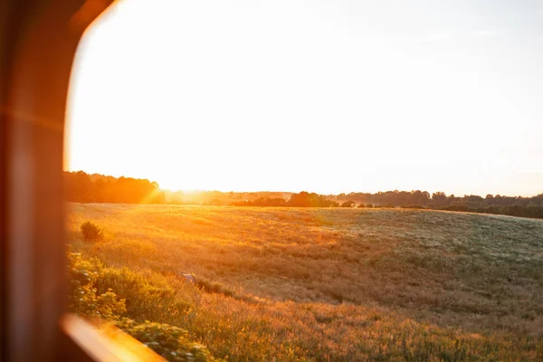 Bellissimo paesaggio con un tramonto arancione brillante e un campo verde fuori dalla finestra del treno. Viaggiare in treno al tramonto . — Foto Stock