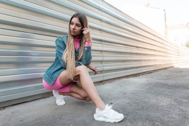 Çekici pembe spor takım elbise beyaz spor ayakkabı şık bir kot ceket sevimli genç kadın sarışın parlak güneşli bir yaz gününde gümüş çit yakınında oturuyor. Amerikalı kız modeli açık havada poz veriyor.