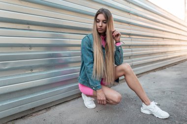 Beyaz trendy spor ayakkabı lar içinde yaz şık kot giysiler sarı uzun saçlı genç güzel kadın bir yaz güneşli bir günde metal bir duvar yakınında bir şehirde oturur. Modern kız dışarıda dinleniyor. Retro tarzı.