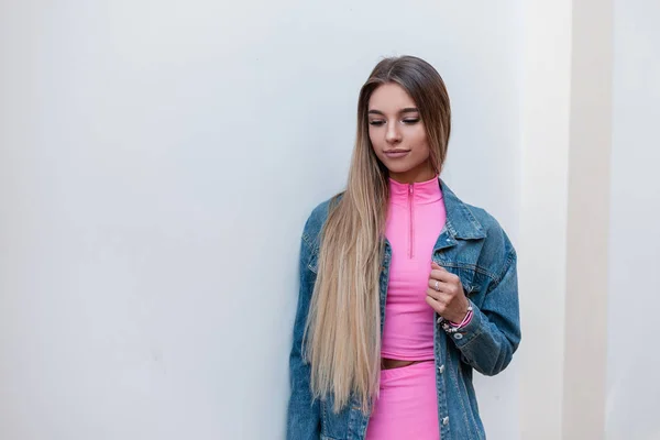 Jolie jeune femme blonde glamour dans un costume sportif d'été rose à la mode dans une veste en jean bleu élégant profitant d'un repos dans la ville. Belle fille mignonne modèle de mode se tient près du mur blanc . — Photo