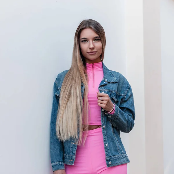 European beautiful young woman in trendy pink top in pink shorts in a denim jacket with blond long hair relaxes standing near a vintage wall in the city. Attractive cute girl outdoors. Youth style. — Stock Photo, Image