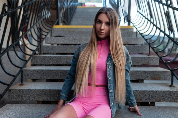 Pretty beautiful young woman with blond long hair in a vintage jeans jacket in a pink top in a stylish pink t-shirt rest on a vintage staircase outdoors in the street. Attractive girl model relaxes.