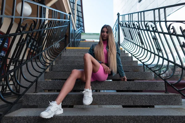 Die glamouröse junge Frau mit langen blonden Haaren in modischer Sommerkleidung in stylischen Turnschuhen entspannt auf Steintreppen in einer Straße der Stadt sitzend. schönes urbanes Mädchen genießt das Wochenende. — Stockfoto