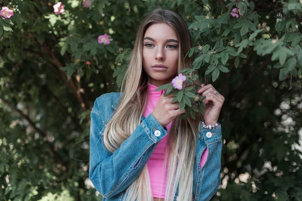 Mulher atraente muito jovem em um top rosa elegante em uma jaqueta de ganga na moda azul de pé goza da natureza perto de um arbusto de floração verde no parque em um dia de verão. Modelo moderno glamoroso da menina .Style . — Fotografia de Stock