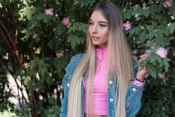 Luxe jonge vrouw met lang haar in een stijlvolle roze top in een blauw denim jasje ontspant staande in de buurt van een struik met bloemen in de tuin. Mooie modieuze meisje rust buitenshuis in het Park. — Stockfoto