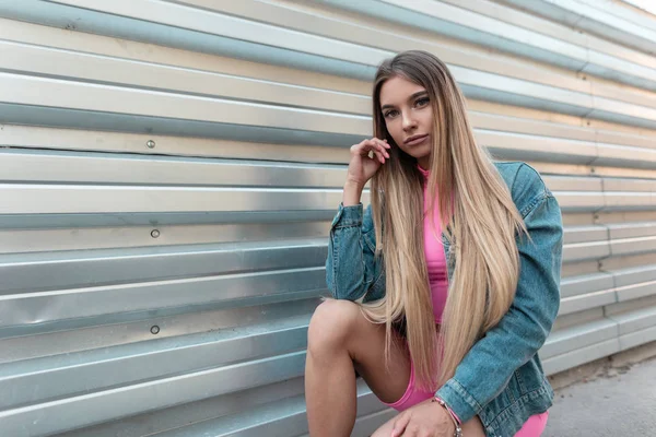 Mulher jovem urbana glamourosa com cabelos longos em um elegante terno esportivo rosa de verão em uma jaqueta jeans na moda azul posando sentado perto de uma parede de metal na cidade. Menina bonita atraente. Estilo de verão . — Fotografia de Stock