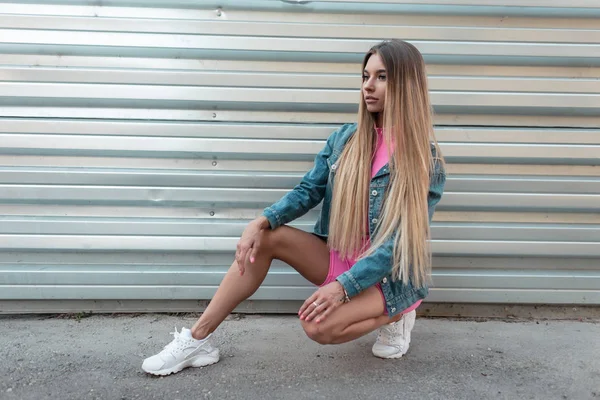 Roztomilá moderní mladá žena blondýnka v módní džínové bundě v módních bílých teniskách ve stylovém růžovém sportovním obleku sedí u stříbrné zdi v letním dnu. Dívčí model Glamour se rozkládá venku. — Stock fotografie
