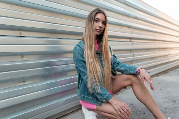 Štíhlá Evropská mladá žena blondýnka v elegantním modrém džínovou kabátku ve stylových růžových trenýrkách sedí u stříbrné zdi na ulici. Krásná dívka si užívá letního slunného dne. Moderní Pouliční móda. — Stock fotografie