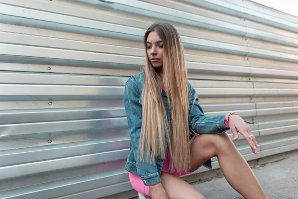 Krásná mladá žena s dlouhými vlasy ve stylové letní růžové sportovní kombinéze v modrém značkové bundě, která sedí u kovového plotu ve městě. Hezká krásná dívka odpočívá. Letní styl. — Stock fotografie