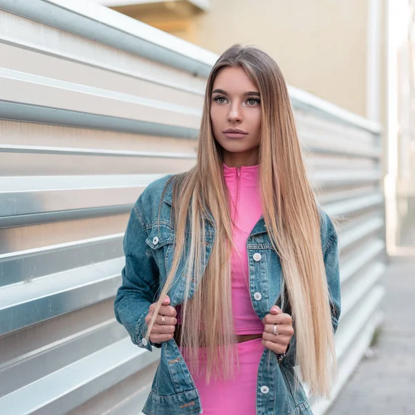 Urocza, efektowna młoda kobieta z blond długimi włosami w niebieskiej stylowej marynarkę denim w modnej różowy top w modne różowe szorty stwarza w pobliżu Srebrnego ogrodzenia. Modne ładne dziewczyny w mieście. Styl letni. — Zdjęcie stockowe