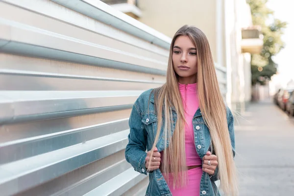 Moderna mujer atractiva rubia con el pelo largo en pantalones cortos elegantes de color rosa en una parte superior de color rosa en una chaqueta de mezclilla de moda se encuentra en la ciudad cerca de una pared vintage en un día de verano. Hermosa chica linda modelo — Foto de Stock