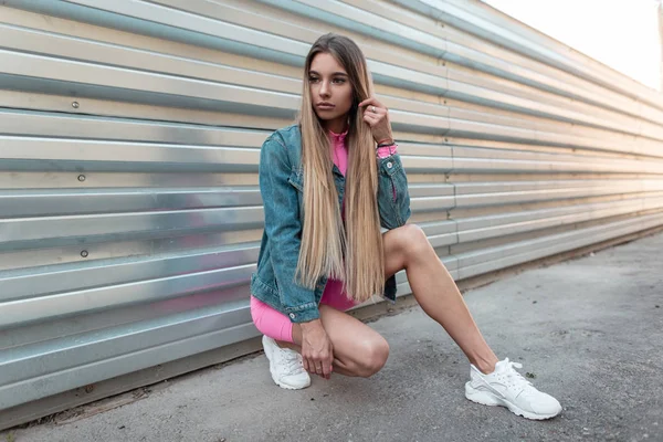 Jeune belle femme aux cheveux longs blonds en été vêtements élégants en denim blanc baskets tendance se trouve dans une ville près d'un mur de métal par une journée ensoleillée d'été. Fille moderne se reposant à l'extérieur. Style rétro . — Photo