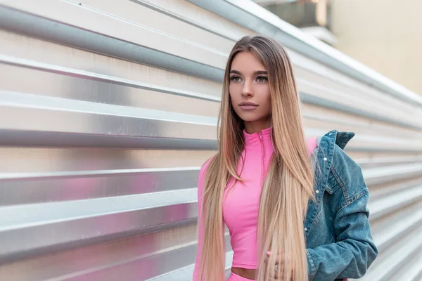 아름다운 긴 금발 머리와 세련된 핑크 티셔츠에 세련된 데님 재킷에 고급스러운 도시 젊은 여성은 금속 벽 근처 야외 포즈. 매혹적인 소녀는 여름 날에 산책을 즐긴다.. — 스톡 사진
