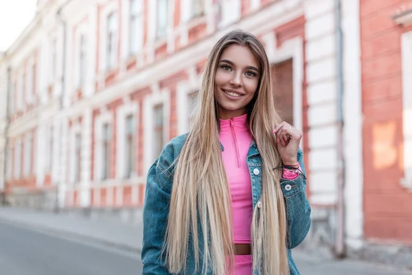Ευτυχισμένη αστική νεαρή γυναίκα με θετικό χαμόγελο με ξανθά μακριά μαλλιά σε ένα μοντέρνο σακάκι τζιν σε ένα κομψό ροζ κορυφή ποζάρει κοντά σε ένα vintage λευκό κτίριο στο δρόμο. Πολύ χαρούμενο κορίτσι περπατάει στην πόλη. — Φωτογραφία Αρχείου