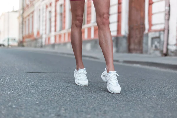 Sportieve jonge vrouw met slanke gelooide benen in stijlvolle witte sneakers loopt langs de straat. Modieuze Damesschoenen. Zomer stijl. Close-up van vrouwelijke benen met schoenen. — Stockfoto