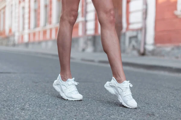 Moderne jonge vrouw met slanke mooie benen in modieuze witte sneakers loopt langs de straat. Stijlvolle sportieve Damesschoenen. Zomer stijl. Close-up van vrouwelijke benen met schoenen. — Stockfoto