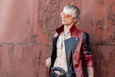 Bir vintage silah ile kot çıplak gövde kırmızı trendy gözlük moda deri siyah-kırmızı ceket güzel seksi genç hipster adam açık havada metal duvar yakınında pozlar. Yakışıklı adam.