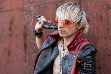 Eski paslı bir binanın yakınında poz kırmızı gözlüklü şık kırmızı-siyah deri ceket çıplak gövde ile Serin genç sarışın adam. Klasik silahı olan yakışıklı seksi hipster adam. Geleceğin punk tarzı