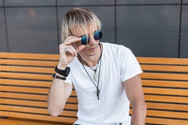 Vintage yuvarlak güneş gözlüğü modaya uygun bir t-shirt şık saç modeli ile Yakışıklı genç adam sarışın şehirde ahşap bir bankta oturan dinleniyor. Avrupalı adam hipster açık havada rahatlatır. Yaz stili.